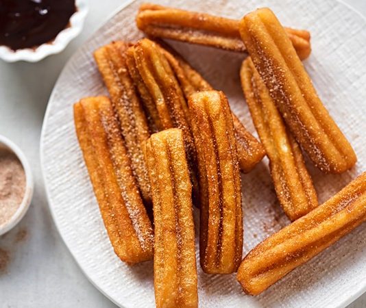Churros: Sokak Lezzetinden Sofralara Uzanan Tatlı Hikayesi Churros, İspanya kökenli, kızartılarak hazırlanan tatlı hamur çubuklarıdır. 19. yüzyılda İspanya’da ortaya çıkan bu lezzet, kısa sürede Latin Amerika ve dünyanın farklı bölgelerine yayılmıştır. Geleneksel olarak kahvaltıda veya akşamüstü çay saatlerinde tüketilen churros, sıcak çikolata veya karamel sosla birlikte sunulur. Konulu bir haber görseli.