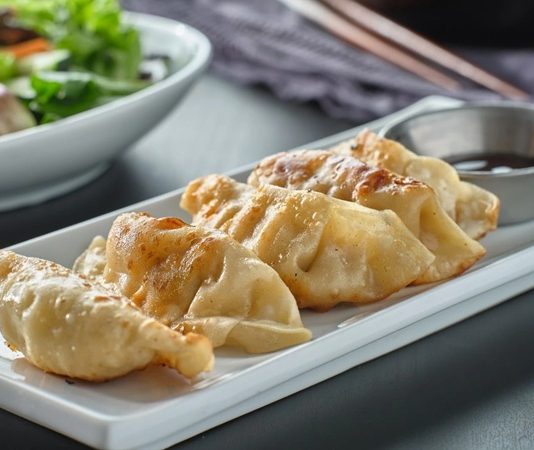 Gyoza, Japon mutfağının en sevilen yemeklerinden biridir ve kökeni Çin’e dayanmaktadır. Aslında Çin’deki jiaozi adı verilen hamur işinden esinlenerek Japonya’ya taşınmış ve zamanla farklı bir kimlik kazanmıştır. Japon mutfağına özgü pişirme teknikleri, gyozayı benzersiz bir hale getirmiştir. Bugün hem Japonya’da hem de dünyanın birçok ülkesinde gyoza, restoranların ve ev sofralarının vazgeçilmez lezzetleri arasında yer almaktadır. Konulu bir haber görseli.