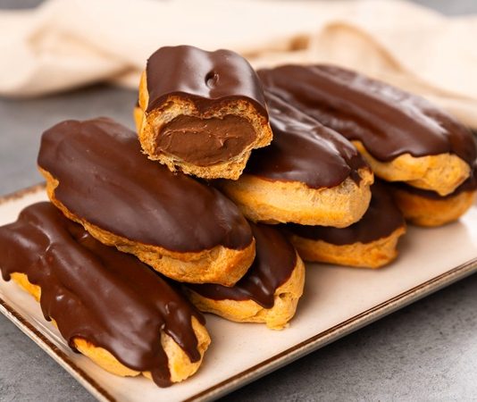 Eclair’in Tatlı Dünyasına Yolculuk Fransa’dan Dünyaya Uzanan Eşsiz Bir Lezzet Fransız pastacılığının dünya mutfağına kazandırdığı en özel tatlardan biri olan eclair, yalnızca görünümüyle değil, ince dokusu ve hafif dolgusu ile de tatlı severlerin vazgeçilmezi olarak gösteriliyor. Günümüzde pek çok pastane menüsünde rastladığımız bu tatlı, aslında aristokrat mutfaklarından çıkıp modern mutfaklara taşınan çok katmanlı bir tarihe sahip. Özellikle çikolata kaplaması ve iç dolgusu ile bilinen eclair, Avrupa’da uzun yıllardır tüketilen bir tatlı olmakla birlikte, son yıllarda Türkiye’de de giderek daha fazla tercih edilmeye başladı. Pastacılığın gelişimi ile beraber farklı dolgular, farklı çikolata türleri ve daha çeşitli glazürlerle sunulan bu tatlı, gastronomik açıdan ciddi bir çeşitliliğin de öncüsü haline geldi. Konulu bir haber görseli.