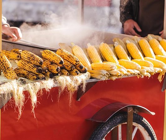 Sokak Satıcıları Kültürü Şehirlerin Hafızasında Yaşayan Lezzetler Şehir hayatının en canlı unsurlarından biri olan sokak satıcıları, yalnızca hızlı ve ulaşılabilir yiyecekler sunan kişiler değil, aynı zamanda kentsel kültürün taşıyıcıları olarak görülüyor. Dar sokaklardan geniş meydanlara kadar uzanan bu hareketli dünya, geçmişten bugüne toplumların yeme içme alışkanlıklarını, ekonomik yapısını ve sosyal ilişkilerini gözler önüne seriyor. Günümüzde modern restoran zincirlerinin artmasına rağmen sokak satıcıları kültürü, samimiyeti ve özgünlüğüyle varlığını korumaya devam ediyor. Konulu bir haber görseli.
